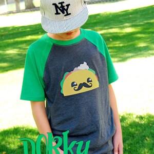 May include: A child wearing a green and gray baseball tee shirt with a cartoon taco graphic. The shirt has the text "Dorky prints n' doodles www.dorkydoodles.com" on the bottom. The child is wearing a striped baseball cap with the letter "NY" on the front.