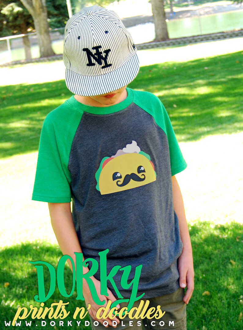 May include: A child wearing a green and gray baseball tee shirt with a cartoon taco graphic. The shirt has the text "Dorky prints n' doodles www.dorkydoodles.com" on the bottom. The child is wearing a striped baseball cap with the letter "NY" on the front.