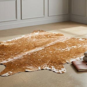Op de afbeelding: Een bruin en wit koeienhuidkleed met een onregelmatige organische vorm, voorzien van een gevlekt patroon van licht- en donkerbruine tinten met witte spikkels, vergezeld door een stapel gevouwen textiel in neutrale tinten en een bruin lederen notitieboek.