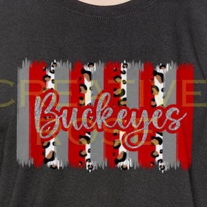 May include: A charcoal gray short-sleeve t-shirt featuring a central graphic design of vertical red, gray, and leopard print brushstrokes. The word Buckeyes is written across the center in a red cursive font with a glittery silver outline.