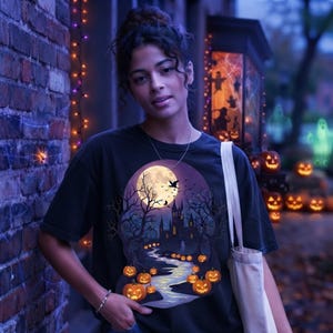 Op de afbeelding: Een zwart t-shirt met een grafische print van een volle maan, een donker kasteel, kale bomen en een kronkelend pad verlicht door gloeiende oranje pompoenen. Het ontwerp bevat silhouetten van vliegende vogels tegen de nachtelijke hemel.
