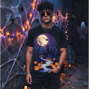 Może przedstawiać: Czarny t-shirt z krótkim rękawem z nadrukiem przedstawiającym nawiedzony zamek w świetle pełni księżyca, otoczony bezlistnymi drzewami i krętą ścieżką wyłożoną świecącymi pomarańczowymi dyniami na tle ciemnofioletowego i niebieskiego nieba.