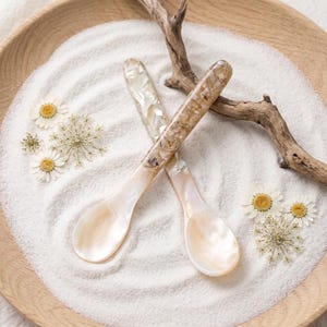 May include: Two spoons with iridescent, pearlescent bowls and handles embedded with crushed shell fragments in shades of beige, cream, and white. The spoons are arranged in a cross shape, showcasing a smooth, glossy finish and natural textures.