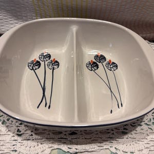 May include: An oval-shaped white ceramic serving dish with a central divider. The interior features two identical hand-painted floral motifs in dark blue and orange. The rim of the dish is accented with a thin dark blue line, providing a clean finish.