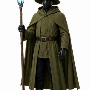 Può includere: Un mantello con cappuccio verde oliva indossato sopra una tunica abbinata e pantaloni neri. L'outfit include un cappello a punta con un emblema a mezzaluna, una cintura in pelle marrone con fibbia in metallo, stivali marroni e un bastone di legno.