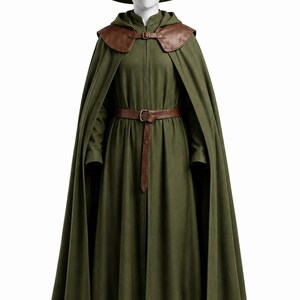 Può includere: Un mantello con cappuccio verde oliva e un cappello a punta abbinato indossati sopra un lungo abito verde oliva. L'insieme presenta una mantellina da spalla in pelle marrone con chiusura a fibbia e una cintura in pelle marrone abbinata in vita, con stivali marroni.