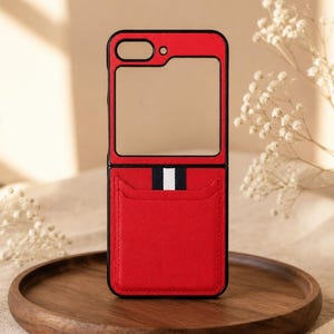 Op de afbeelding: Een rood opvouwbaar telefoonhoesje met een zwart frame en een rechthoekige uitsparing voor de cameralens. Het onderste gedeelte is voorzien van een kaartvak met een verticaal wit en zwart gestreept accent op de bovenrand van het vak.