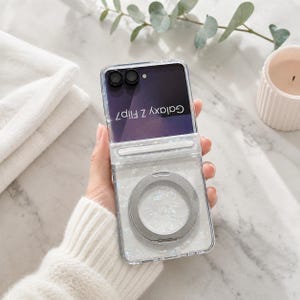 May include: A clear protective phone case for a Galaxy Z Flip7, featuring a white pearlescent shell-like inlay on the bottom half and a circular silver magnetic ring mount centered on the back, with the phone screen visible at the top.