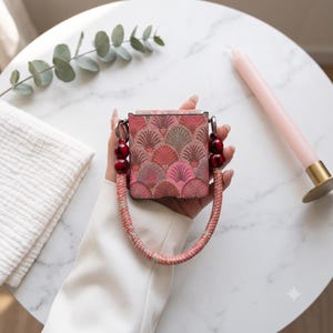 Peut inclure: Un étui de protection carré orné d'un motif répétitif en forme d'éventail dans des tons de rose, rouge et doré. L'étui comprend une anse circulaire en cordon tressé multicolore, agrémentée de deux grosses perles marbrées rouge foncé.