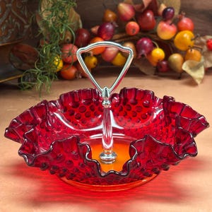 May include: A deep red glass serving dish featuring a hobnail texture and a ruffled, scalloped edge. The dish includes a central silver-toned metal handle with a triangular top loop, designed for carrying the bowl by its center post.