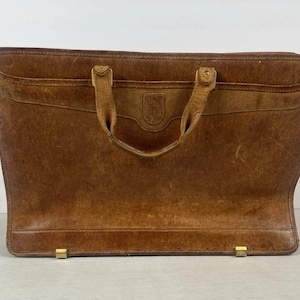 May include: A rectangular brown leather briefcase featuring a weathered texture, two top handles attached with metal hardware, a horizontal stitched panel across the upper section, and two small metal feet at the base for stability.