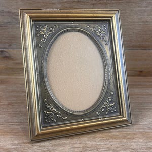 May include: A rectangular picture frame with a gold-toned outer border and a dark, metallic-finished inner panel. The inner panel features an oval center opening surrounded by embossed floral scrollwork details in each of the four corners.