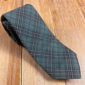 May include: A necktie featuring a dark forest green base colour with a classic plaid pattern consisting of thin, intersecting black and white lines. The fabric has a matte finish and is folded over itself at the top, showing a pointed bottom end.