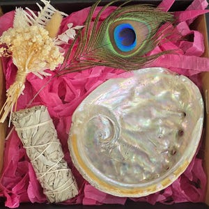 May include: A gift box filled with bright pink shredded paper containing an iridescent abalone shell, a bundle of white sage tied with string, a peacock feather with blue and green eye detail, and a small bouquet of dried cream-coloured flowers.