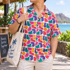 Puede incluir: Una camisa de manga corta con botones y un patrón repetitivo de gatos en amarillo, rosa, morado, rojo y verde. Un bolso de tela beige con gráficos de palmeras azules y el texto "Good Vibes Always" se lleva sobre el hombro.