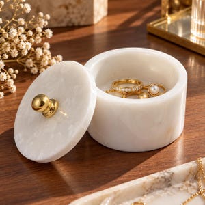 May include: A round white marble jewelry box with a removable lid featuring a polished gold-tone knob. The container holds several gold rings, including one with a small white pearl and another with a band of tiny clear stones set inside.
