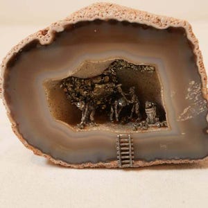 May include: A cross-section of a brown and beige agate geode featuring a hollow center. Inside the geode, small metallic figurines of miners are depicted working with tools, alongside a tiny metal ladder attached to the bottom edge.