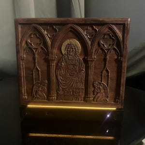 May include: A rectangular wooden panel featuring intricate relief carvings of a central figure with a gold-toned halo, flanked by two kneeling angels within gothic-style arches. The base of the panel is illuminated by a warm light source.