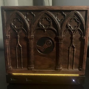 May include: A dark brown wooden computer case featuring intricate gothic arch carvings and quatrefoil details. A circular cutout in the centre displays the Steam logo. The base includes a gold-toned light strip, two USB ports, and a small power button.