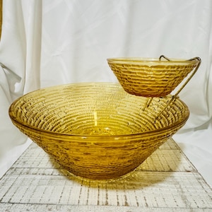 May include: A large amber-colored glass bowl with a textured, concentric ring pattern on the exterior. A smaller matching glass bowl is suspended above the larger bowl by a gold-toned metal bracket attached to the rim of the larger bowl.