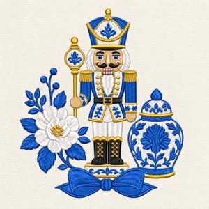 Può includere: Un disegno ricamato raffigurante uno schiaccianoci in uniforme blu e bianca con dettagli dorati, che tiene in mano un bastone. A sinistra, un fiore bianco con foglie e bacche blu. A destra, un vaso con coperchio blu e bianco, sopra un fiocco blu.