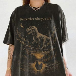 May include: A charcoal gray short-sleeve t-shirt featuring a graphic print of a Tyrannosaurus Rex above a chicken in a landscape. The text "Remember who you are." is printed in a light tan color above the dinosaur, which is set against a starry sky.