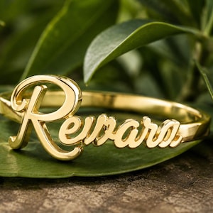 以下が含まれることがあります： 筆記体フォントで「Revara」という名前が刻まれた、光沢のあるゴールドトーンのリング。金属製のバンドは滑らかな円形で、文字がリングの前面部分に組み込まれており、連続したつながりのあるデザインになっています。