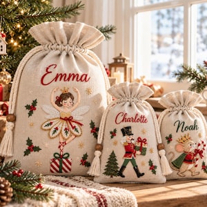 May include: Three beige drawstring gift bags made of a linen-like fabric. The largest bag features the name Emma and a fairy illustration. The medium bag shows the name Charlotte and a nutcracker. The smallest bag displays the name Noah and a mouse.