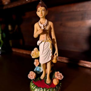 Puede incluir: Una figura de pie de una persona joven con peinado de moño, vistiendo una prenda de tela blanca con un cordón dorado, una bufanda blanca y una marca roja en la frente. La figura sostiene un pequeño recipiente marrón y se apoya sobre una base con flores.