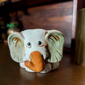 Può includere: Una statuetta di elefante in ceramica con corpo bianco, grandi orecchie verde salvia e una proboscide arancione prominente. L'elefante ha due piccoli occhi neri e una finitura liscia e lucida, raffigurato frontalmente con una forma arrotondata.