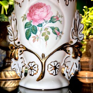 Puede incluir: Un jarrón de cerámica blanca con una ilustración central de una rosa rosa con hojas y capullos verdes. El jarrón está decorado con detalles de hojas doradas en los laterales y motivos florales con bordes dorados en la base, con un borde dorado.