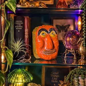 Op de afbeelding: Een handbeschilderd oranje keramieken gezichtssculptuur met opvallende zwarte wenkbrauwen, gele ogen en een brede mond. Het beeld heeft een getextureerd, onregelmatig oppervlak en staat op een glazen plank tussen diverse decoratieve objecten en boeken.