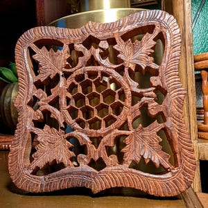 May include: A square wooden trivet featuring an intricate hand-carved design. The center displays a honeycomb pattern surrounded by four detailed maple leaves in each corner. The outer border has a textured, ribbed edge with a dark brown finish.
