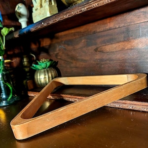 May include: A triangular wooden rack for billiards balls with a smooth, light brown finish. The frame is constructed from solid wood with visible grain patterns, featuring rounded corners and a hollow center designed to hold a set of pool balls.