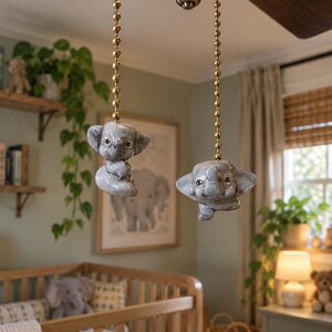 Può includere: Due statuette di elefanti in ceramica grigia attaccate a catenelle a sfere in metallo dorato, progettate come tiranti per ventilatori a soffitto. Ogni elefante presenta una finitura liscia e lucida con dettagli del viso, grandi orecchie e proboscide.