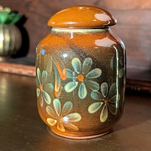 May include: A small, round ceramic jar with a glossy, crackled brown glaze. The body features a repeating pattern of light blue, daisy-like flowers with orange centers. It includes a matching, rounded, removable lid that sits flush on top.