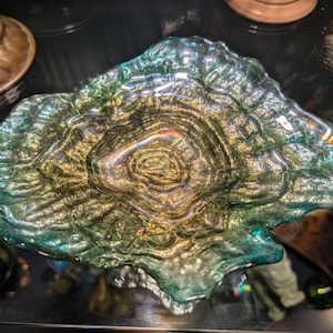 Könnte beinhalten: Eine dekorative Glasschale mit unregelmäßiger, organischer Form und gewellter, strukturierter Oberfläche. Das Glas weist einen Farbverlauf von tiefen blaugrünen Rändern hin zu einer warmen, metallisch goldenen und bernsteinfarbenen Mitte auf.
