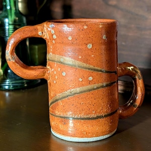 May include: A cylindrical ceramic mug with a speckled orange glaze, featuring two thick, curved handles on opposite sides. The surface is decorated with two dark, diagonal bands that wrap around the body, and the base has a light gray rim.