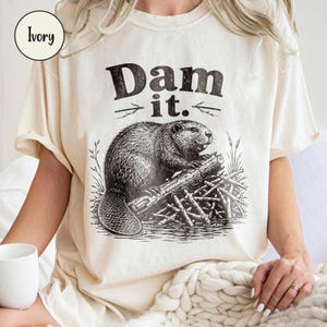 May include: An ivory-coloured t-shirt featuring a black graphic illustration of a beaver perched on a log. The text "Dam it." is printed in a bold, distressed font above the beaver, with small arrow-like symbols pointing outward on each side.
