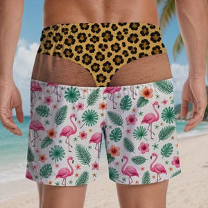 May include: Swim trunks featuring a white base with a repeating pattern of pink flamingos, green palm leaves, and orange hibiscus flowers. The waistband displays a yellow and black leopard print design that creates a visual illusion of a thong.