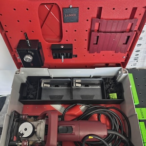 May include: A red and grey Lamello power tool case containing a biscuit joiner with a black power cord. The interior lid features a red pegboard-style organizer with a black L-DOCK label, while the base holds the tool and metal accessories.