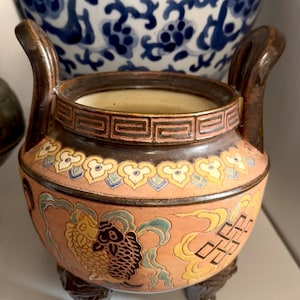 May include: A ceramic, handled pot with a peach-colored exterior, featuring fish and geometric designs in yellow, blue, and brown. The pot has a dark brown rim and handles, and rests on three small feet. A blue and white patterned bowl is in the background.