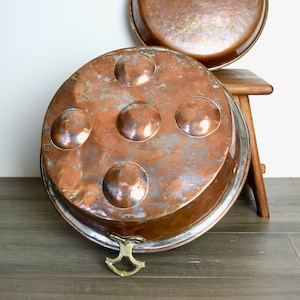 Puede incluir: Dos platos de servir de cobre antiguo con superficies martilladas. El plato más grande tiene un borde liso y el plato más pequeño tiene un borde decorativo con cinco círculos en relieve. Ambos platos tienen un asa de latón.