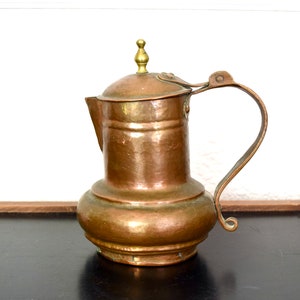 May include: A copper pitcher with a brass lid and handle. The pitcher has a rounded body and a flared spout. The handle is attached to the lid and has a decorative curl at the end.