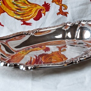 May include: A silver serving dish with an ornate, scalloped edge. The dish is reflecting the image of a colorful tablecloth with a rooster design.