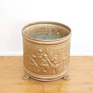 Könnte beinhalten: Ein goldfarbener Metall-Pflanzkübel mit einem geprägten Design von Menschen in einer Szene. Der Kübel hat drei Füße und steht auf einem Holzboden.