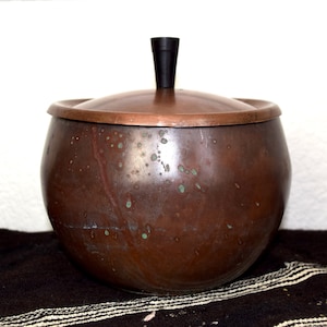 Może przedstawiać: Miedzany garnek z pokrywką i czarnym uchwytem na górze. Garnek ma nakrapiany, postarzony wygląd.