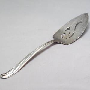 May include: Silver-toned pie server with a decorative, pierced design on the blade. The handle has a curved, elegant shape. The server is designed for serving cakes, pies, or other baked goods.