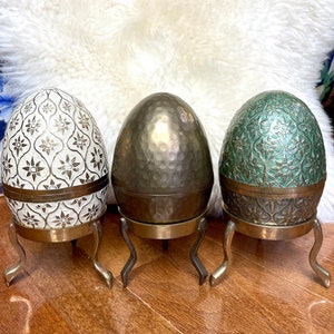 May include: Three decorative egg-shaped boxes with intricate designs. The boxes are made of metal and have a hammered finish. The boxes are on three legs and are arranged in a row.