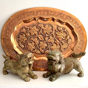 May include: A large, oval copper tray with intricate floral carvings. Two stone Foo Dog statues are in front of the tray. The tray and statues are on a white surface.